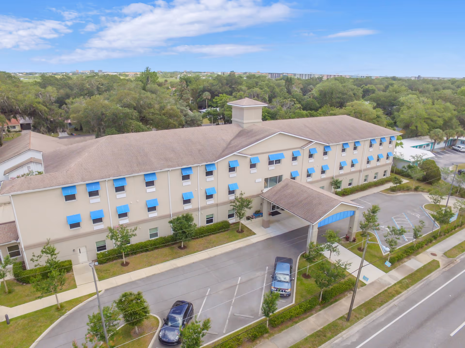 Aerial view of a three-story senior living facility building with beige walls and blue window awnings. The building has a covered entrance with a driveway and parking spaces, surrounded by trees and greenery under a partly cloudy sky.
