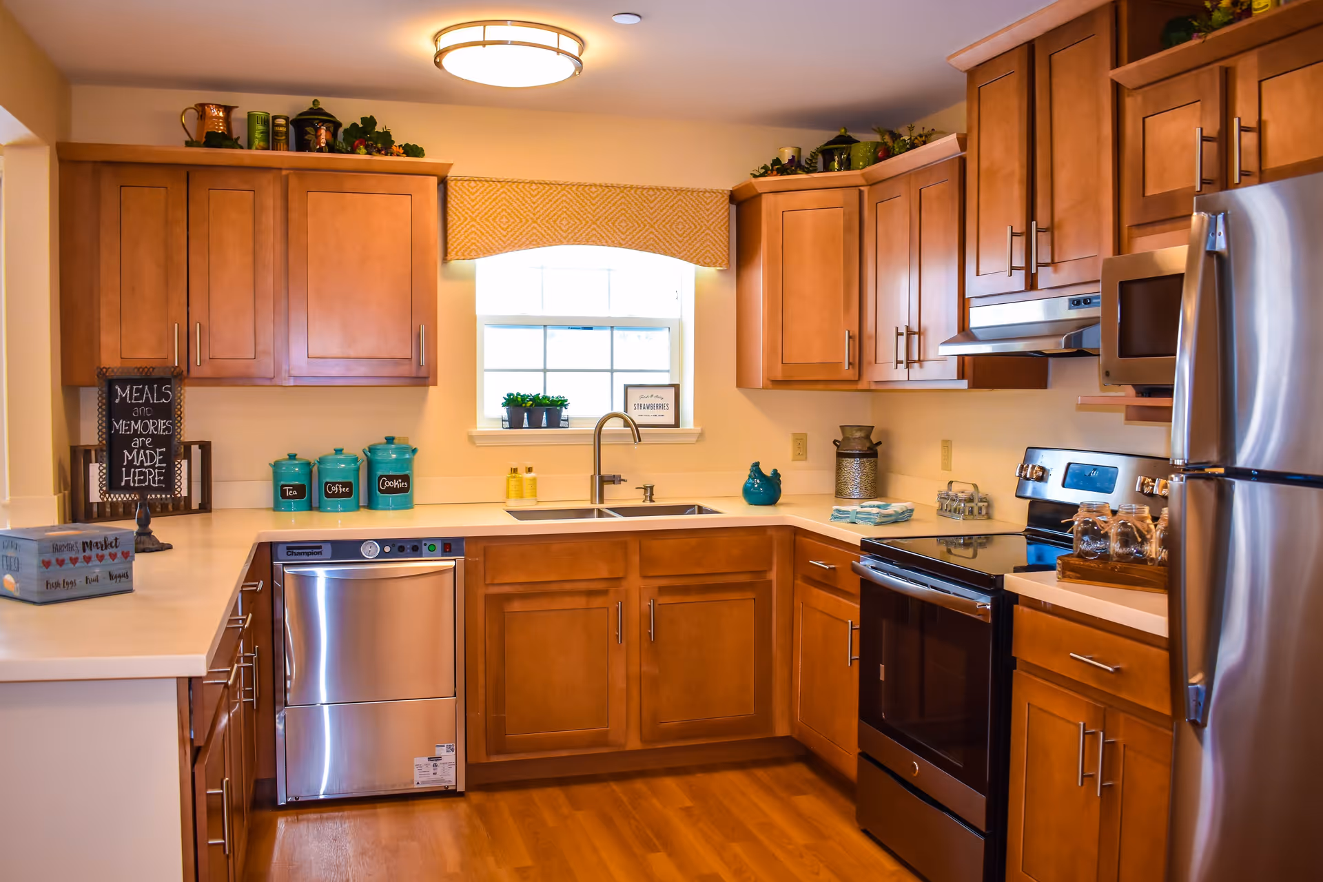 A bright kitchen with wooden cabinets, stainless steel appliances including a dishwasher, oven, microwave, and refrigerator. The countertop has decorative jars labeled Tea, Coffee, and Cookies, a small plant, and a sign that reads 'Meals and Memories are Made Here.' A window with a yellow valance lets in natural light above the sink.