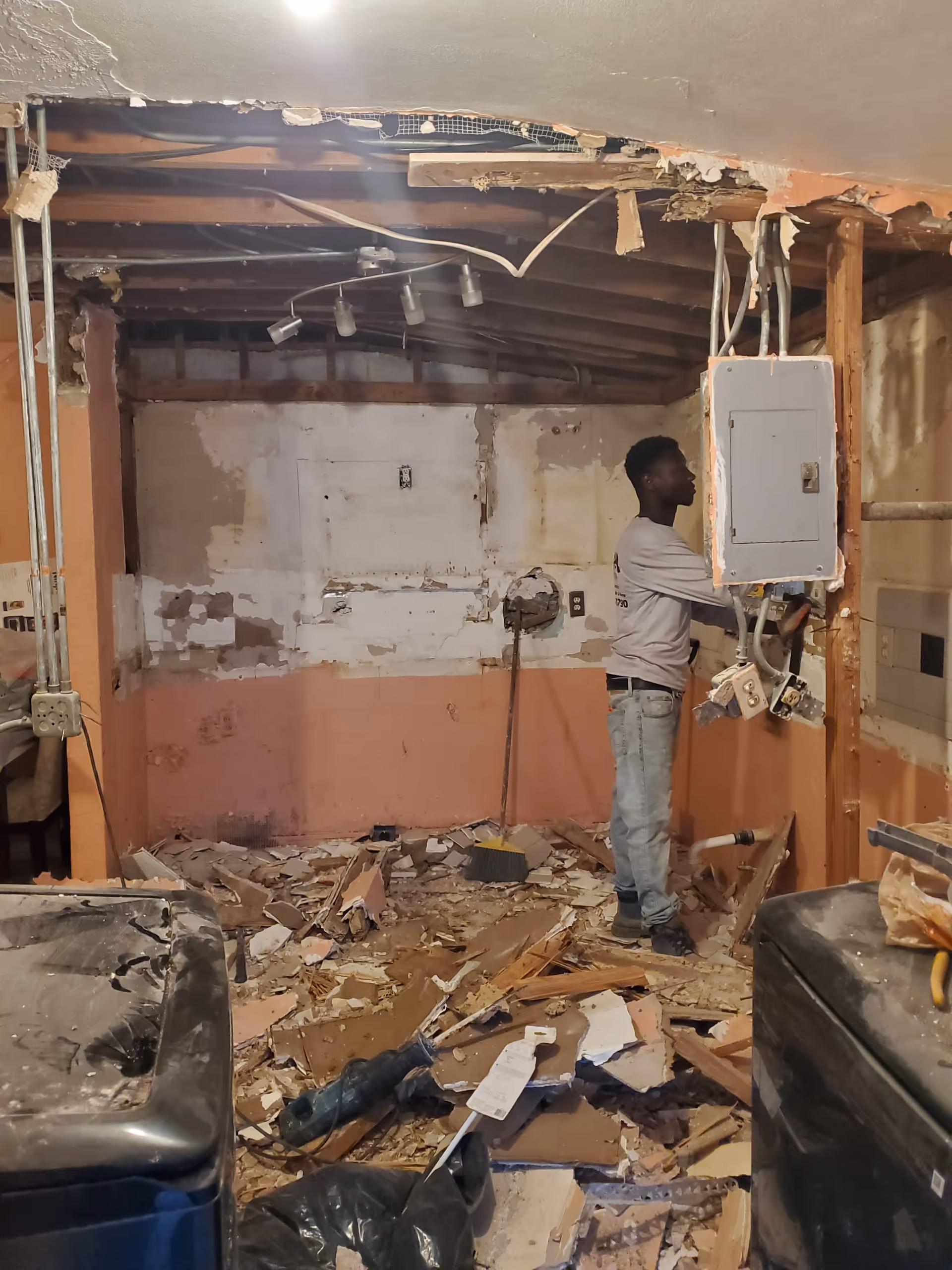 Worker stands amid demolition debris in a damaged unfinished basement with exposed pipes and wiring.