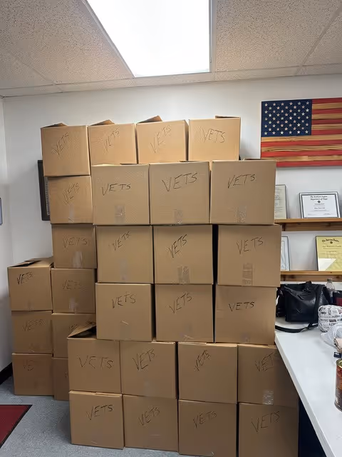 Stacks of cardboard boxes labeled VETS fill a small indoor storage room with an American flag.