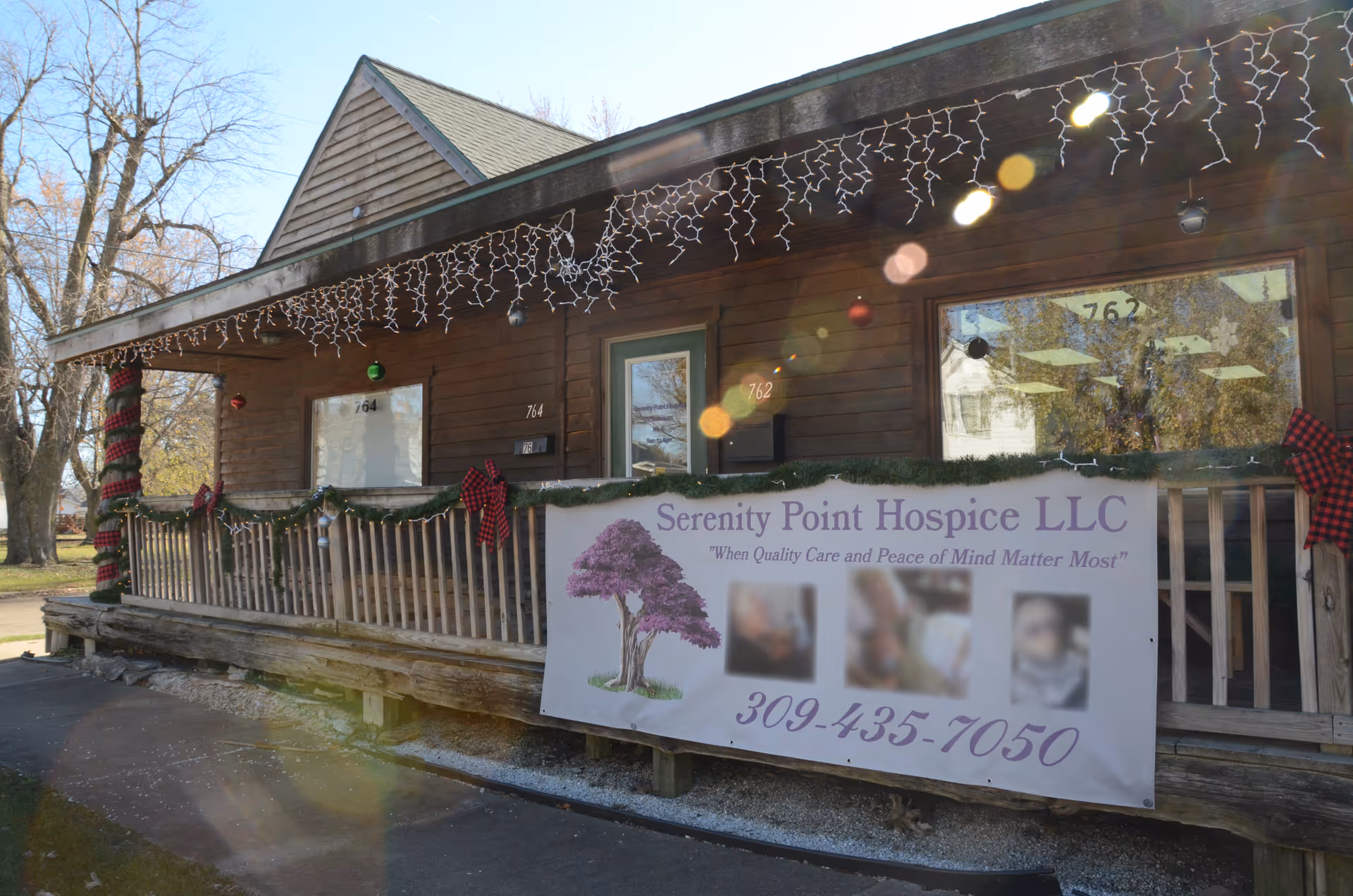 Exterior wooden hospice building with holiday lights and a banner advertising Serenity Point Hospice LLC.