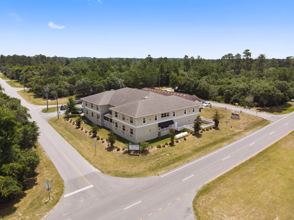 Aerial view of a two-story assisted living facility building named Marion Oaks Assisted Living, surrounded by trees and greenery, located at the intersection of two roads under a clear blue sky.