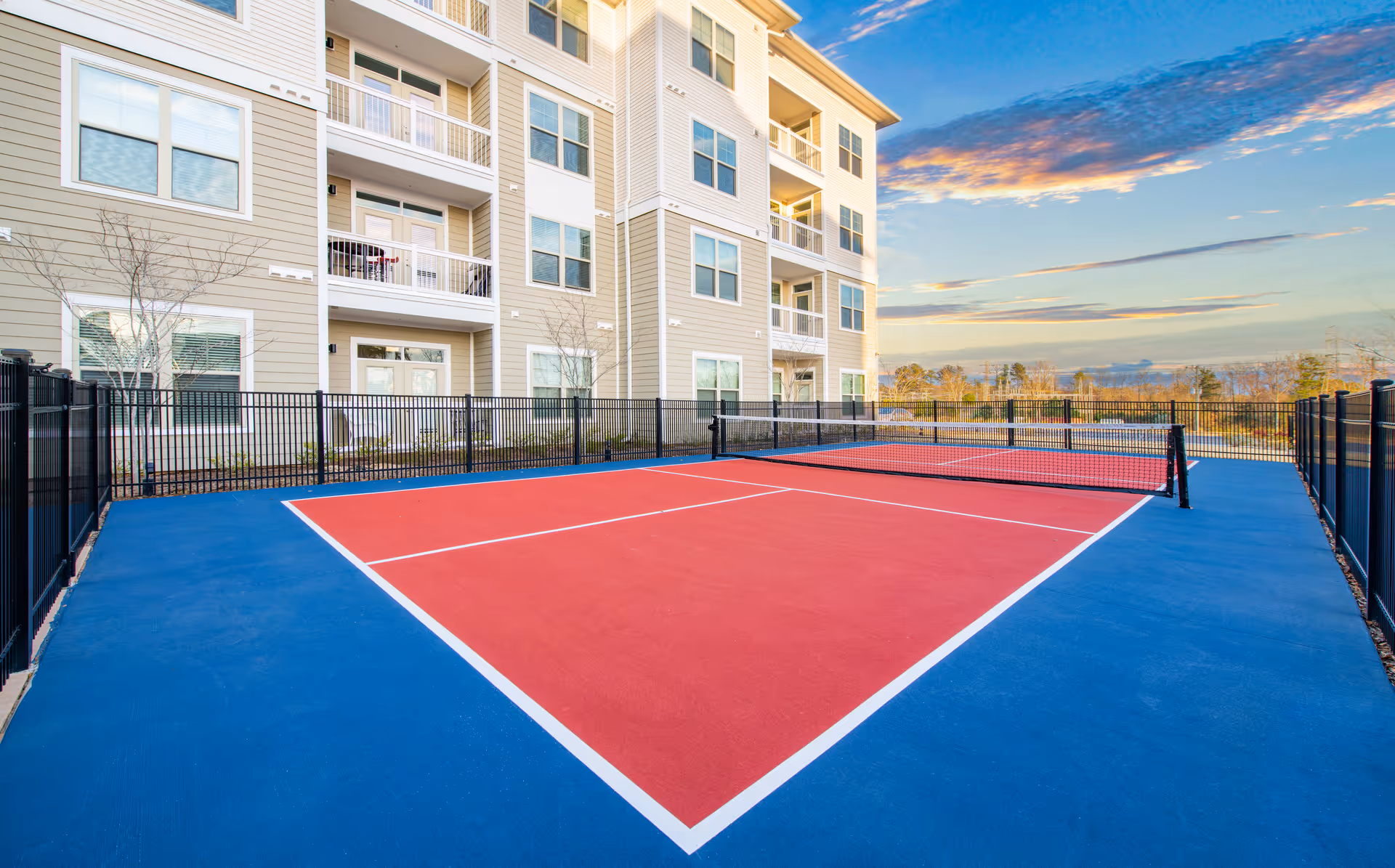 Outdoor pickleball court with a red playing surface and blue surrounding area, enclosed by a black metal fence. In the background, there is a multi-story beige residential building with balconies and windows under a partly cloudy sky during sunset.