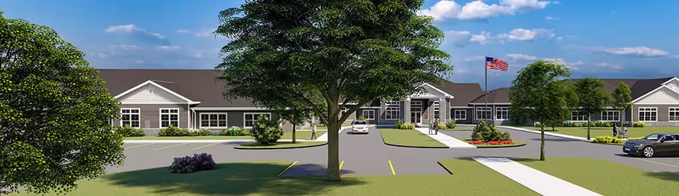 Exterior view of a single-story senior living facility building with a pitched roof, multiple windows, and an American flag on a flagpole in front. The building is surrounded by green lawns, trees, and a parking area with a few cars. The sky is blue with some clouds.