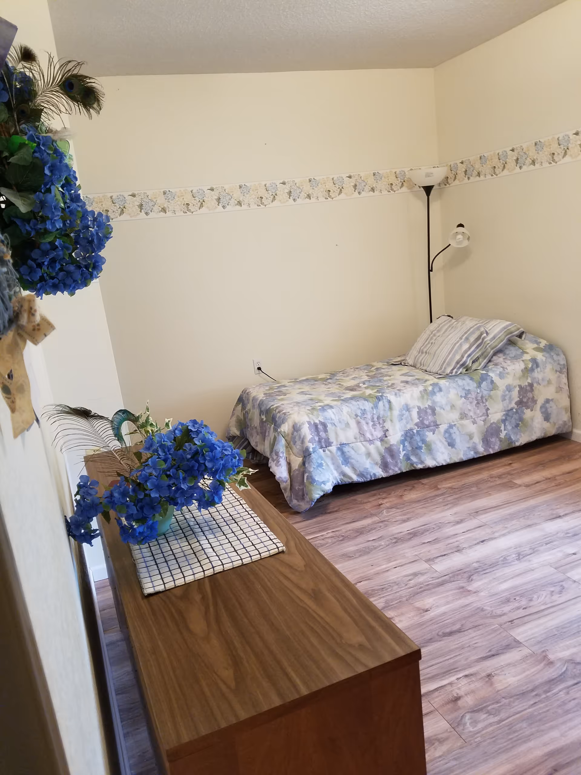 A small senior living bedroom with a floral bedspread, dresser, and blue decor.