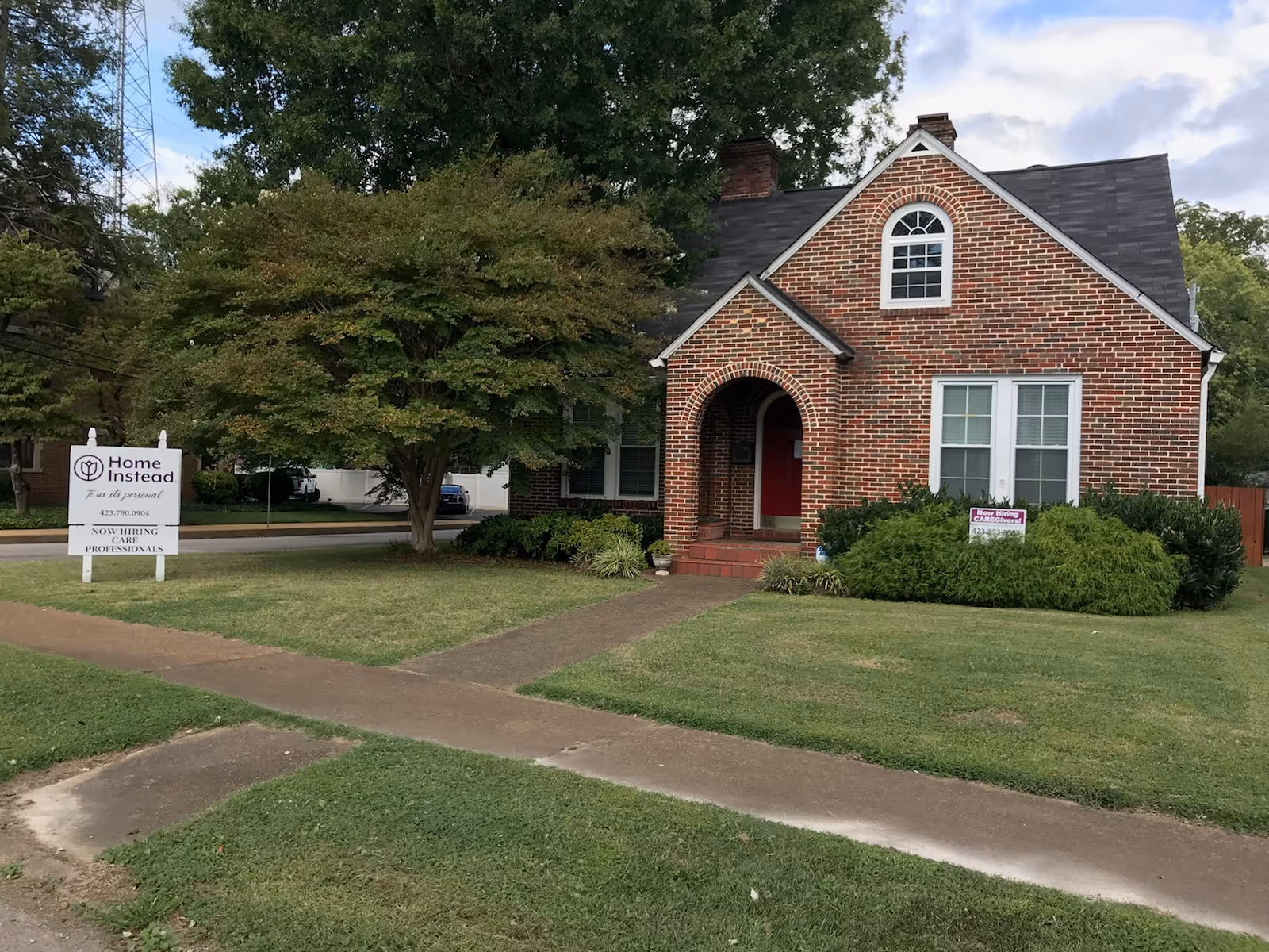 Exterior view of a brick house-style senior care building with lawn, trees, and Home Instead sign.