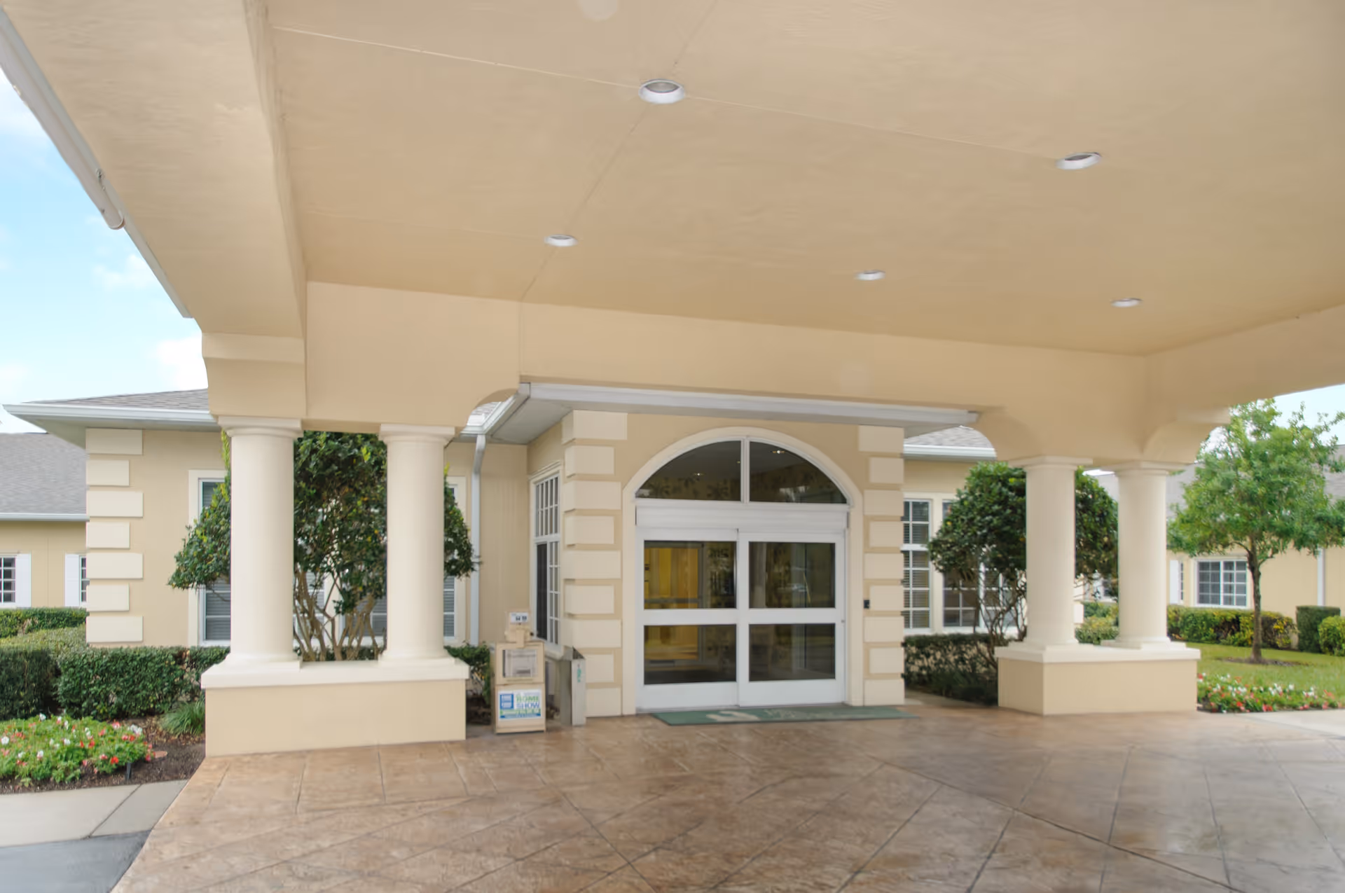 Covered entrance of a beige building with white columns and automatic sliding glass doors, surrounded by neatly trimmed bushes and small trees.