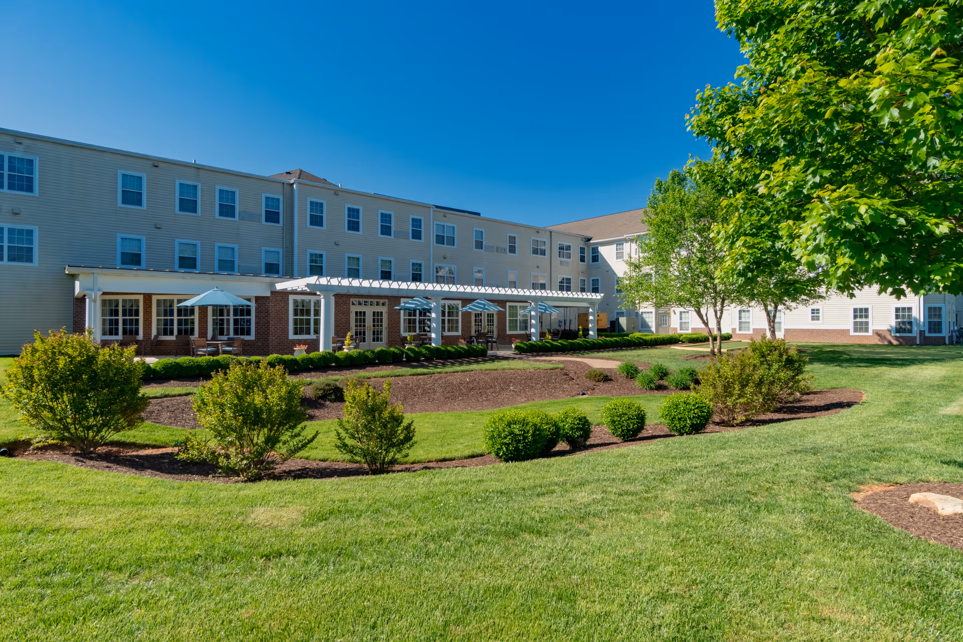Exterior view of a three-story senior living building with a landscaped lawn, patio umbrellas, and trees under a clear blue sky.