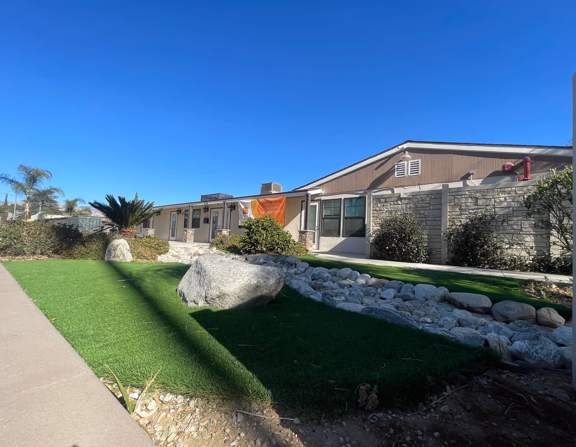 Exterior view of a single-story building with a stone and stucco facade under a clear blue sky. The foreground features a landscaped area with green grass, rocks, and bushes. A sidewalk runs along the left side of the image. There is a banner on the building that reads 'Assisted Living Accepting New Residents'.