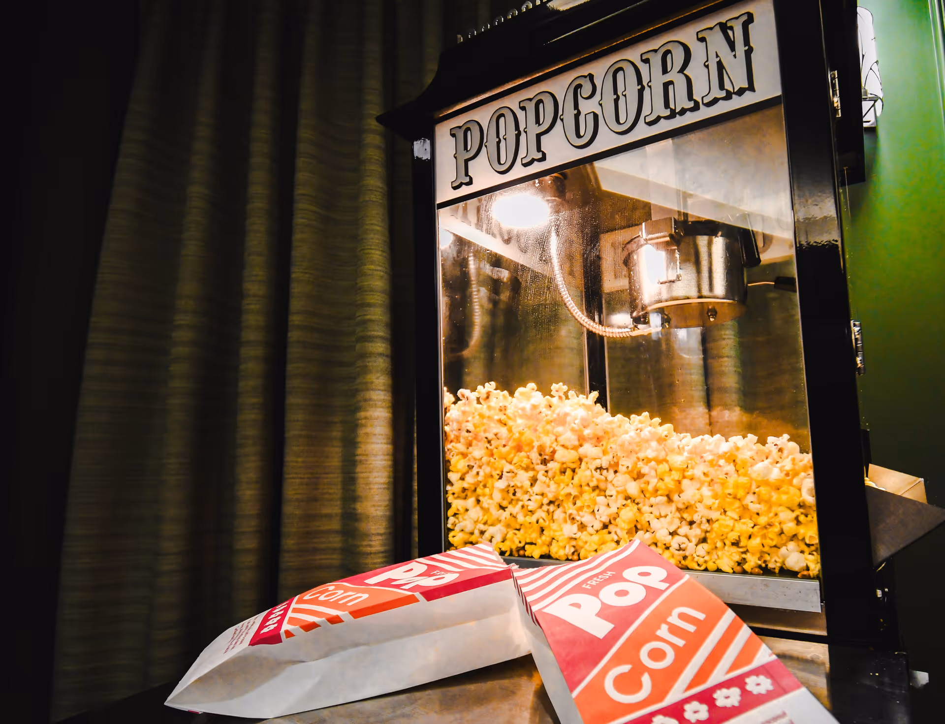 A popcorn machine filled with freshly popped popcorn with two empty red and white striped popcorn bags placed in front of it on a table, set against a backdrop of green curtains.