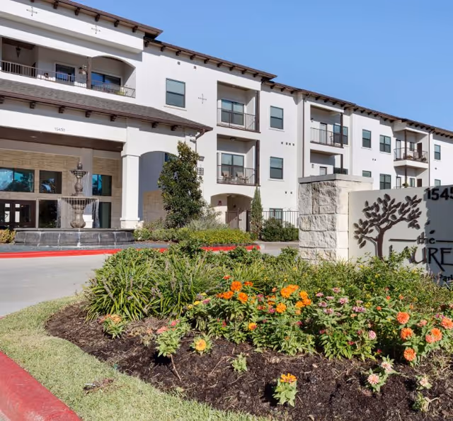 Three-story white senior living building with arched entrance and flower-filled landscaping.