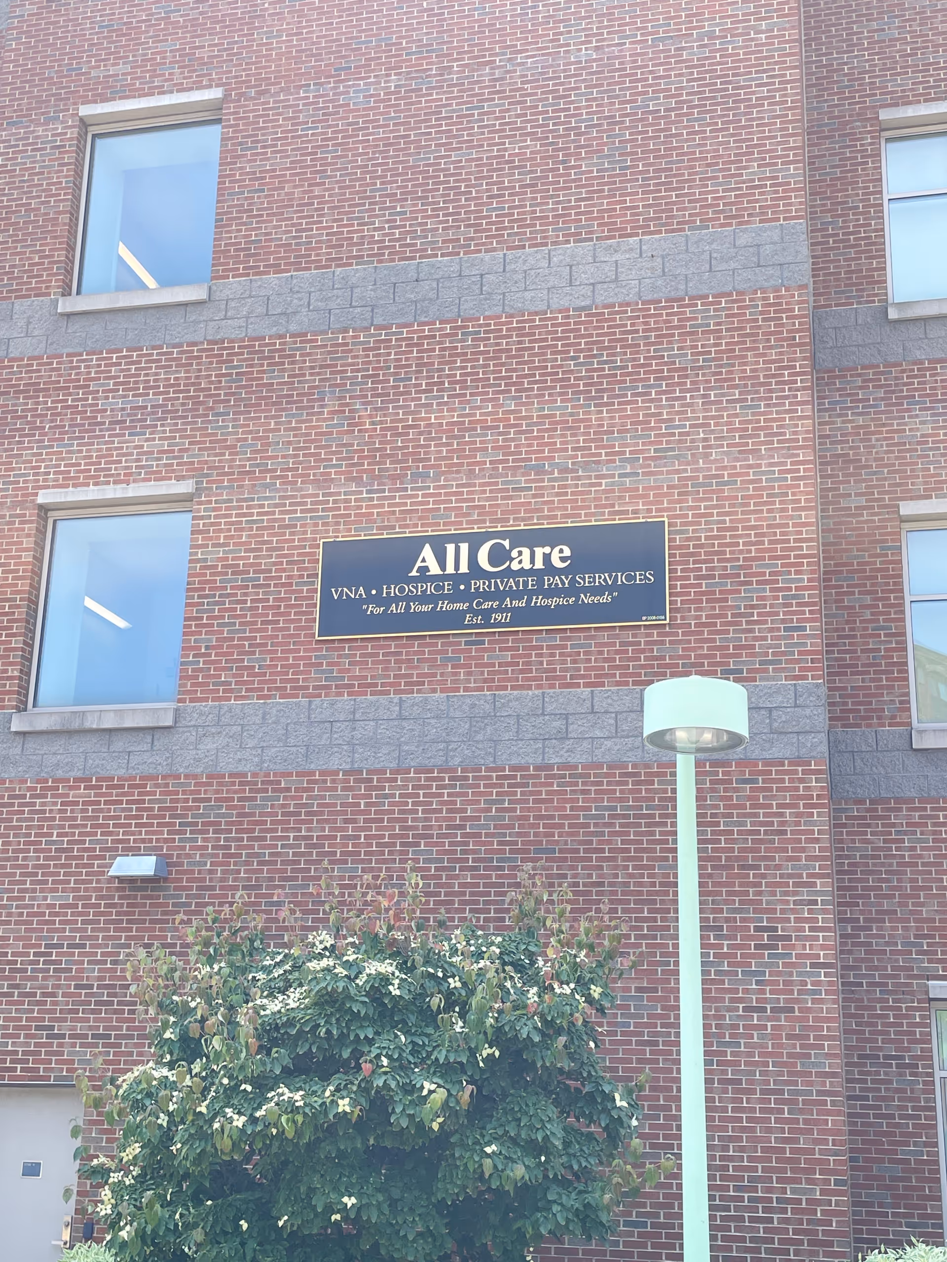Brick building exterior featuring All Care sign, windows, tree, and street lamp.