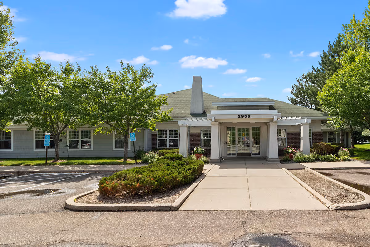 Front exterior view of a single-story senior housing building with a covered entrance, surrounded by green trees and bushes under a blue sky with scattered clouds. The building has a light gray roof and white pillars at the entrance.