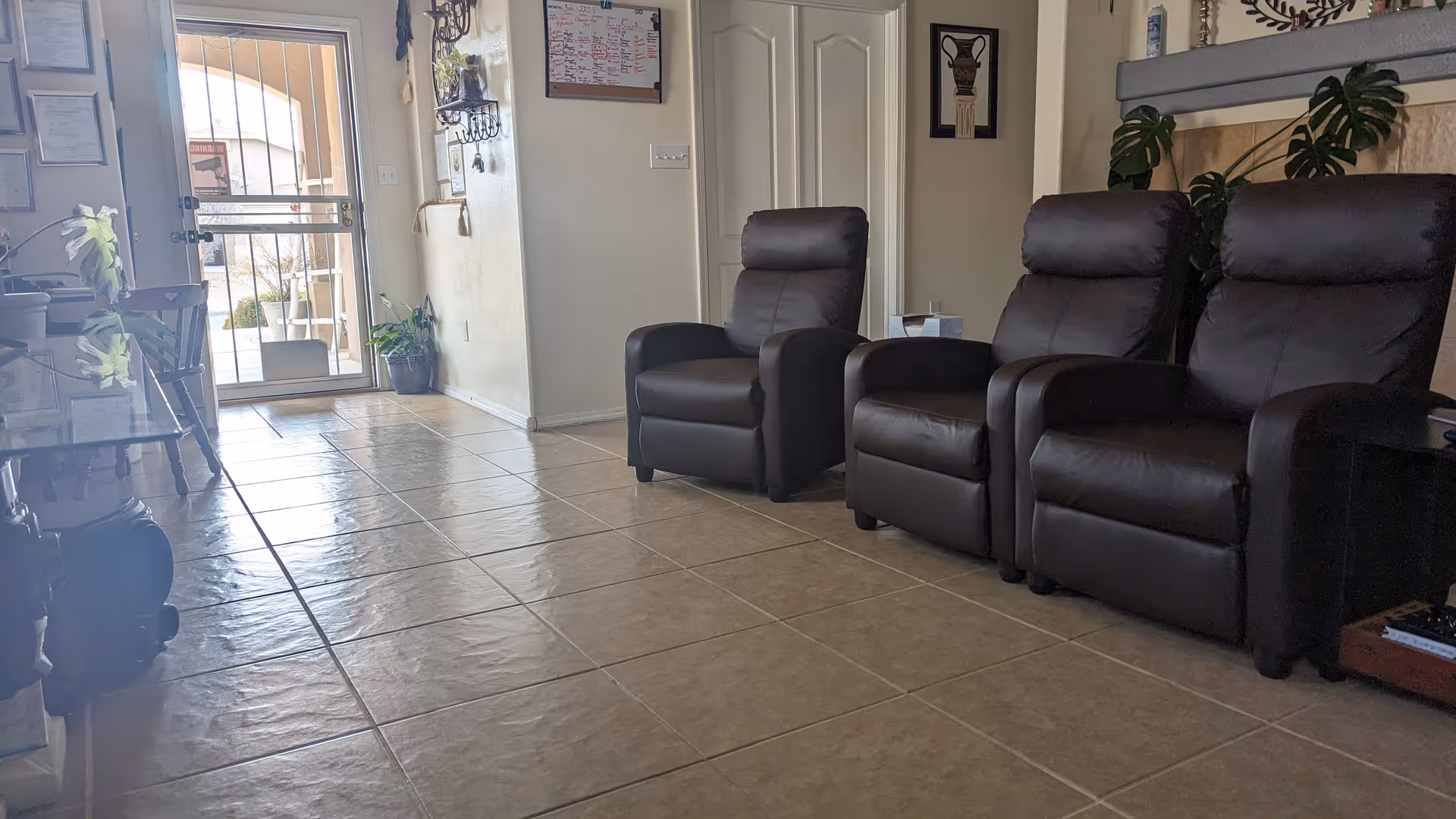 Spacious lounge with brown leather recliners, tiled floor, plants, and a glass door.