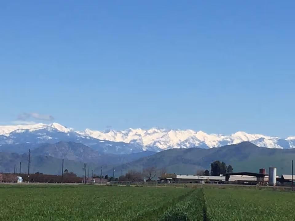 Wide rural landscape with green fields, distant mountains, and a low lying facility building.