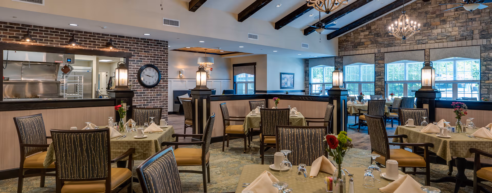 A well-lit dining room in a senior living facility with multiple tables covered in green tablecloths, each set with napkins, glasses, cups, and small flower vases. The room features a combination of brick and stone walls, large windows with blinds, ceiling fans, and decorative lighting fixtures. A kitchen area with stainless steel appliances is visible through a large window opening in the brick wall.