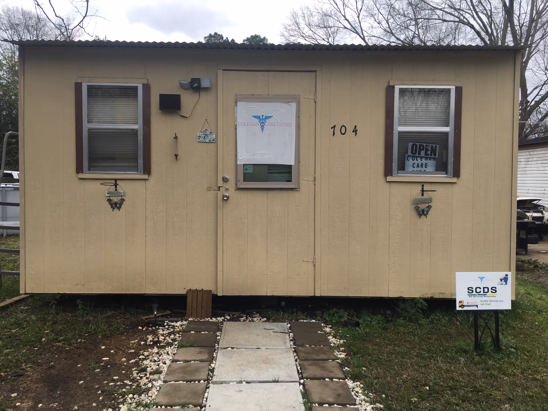 Small beige portable building with two windows, door, and a sign reading Coleman Care Givers.