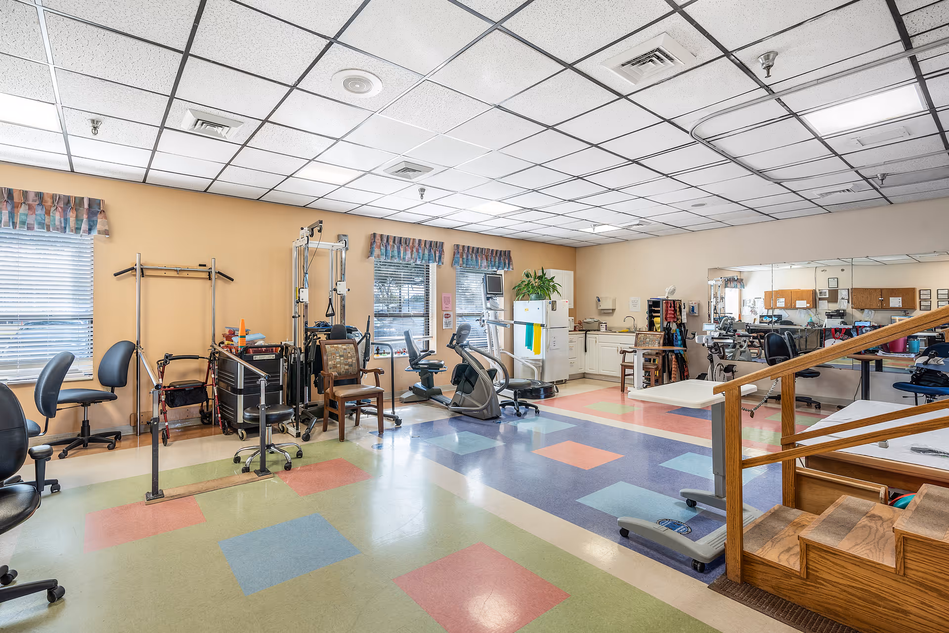 A spacious rehabilitation room with colorful tiled flooring, various exercise and therapy equipment, chairs, a refrigerator, and a large mirror on one wall. The room has beige walls, windows with valances, and a wooden handrail with steps leading to a raised platform.