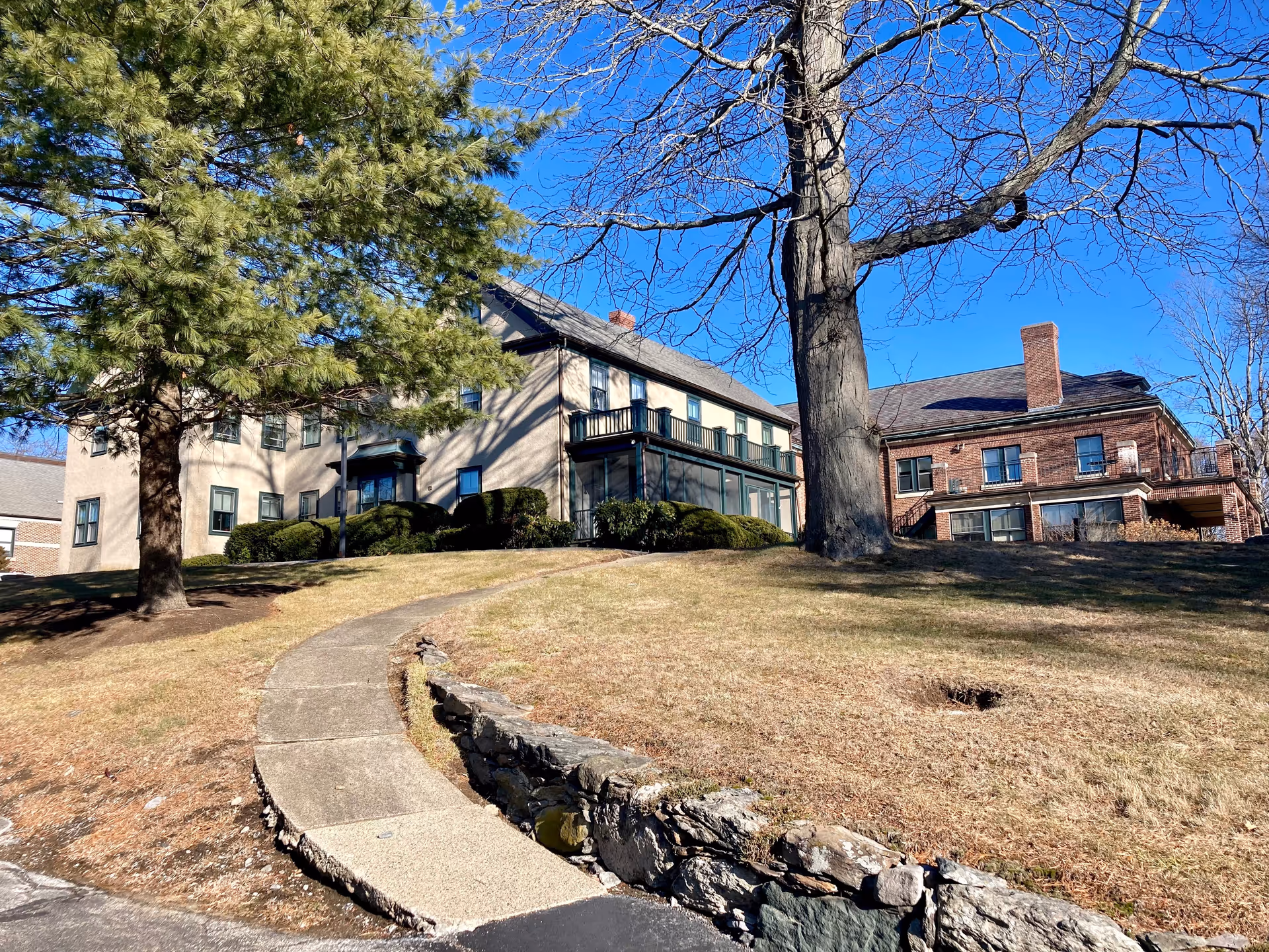 Two connected senior living buildings on a grassy hill with a curved concrete walkway, large trees, and a clear blue sky.