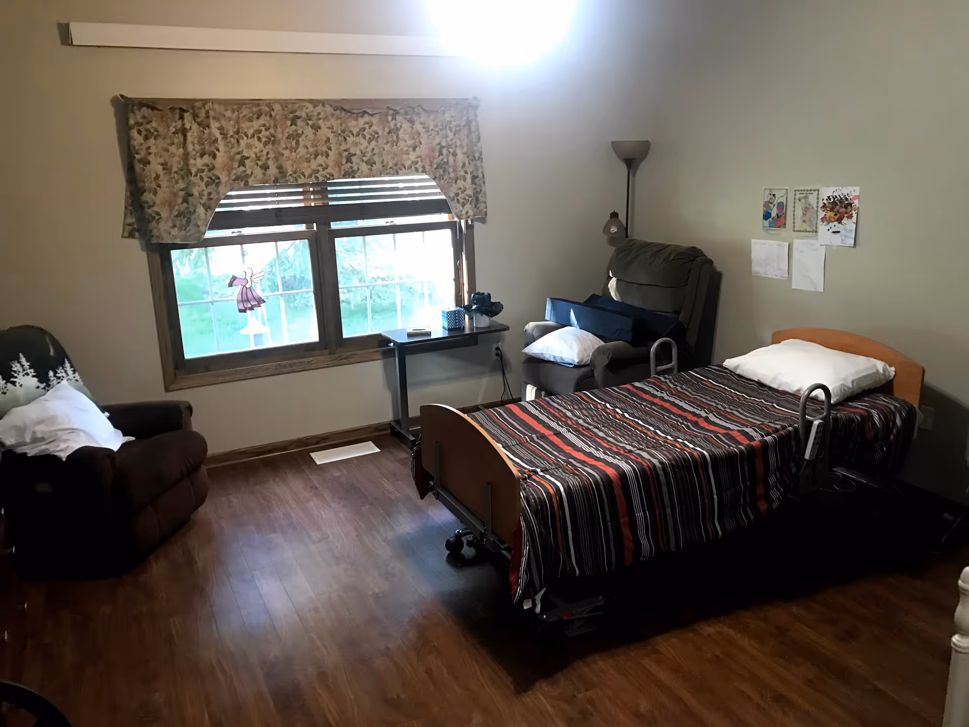 A residential patient room with a single bed, striped bedspread, recliner, window, and wood floors.