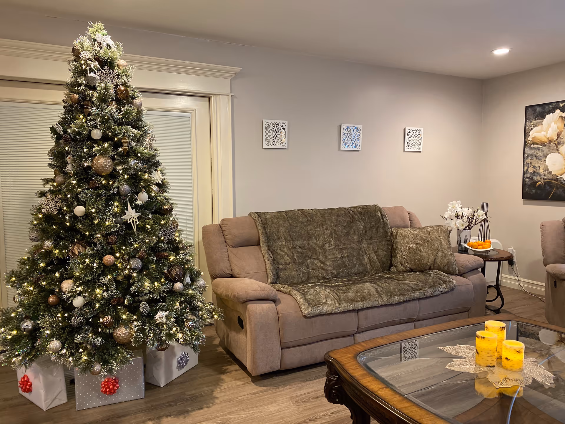 Living room with a decorated Christmas tree and wrapped gifts next to a beige reclining sofa draped with a throw and a coffee table holding candles.
