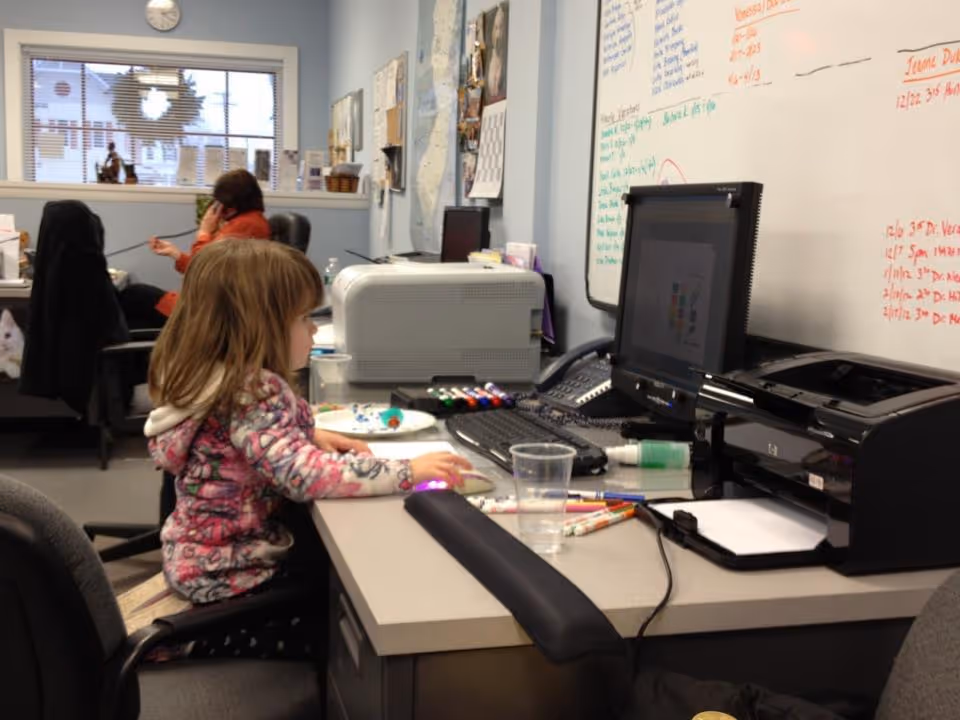 Child at a desk in a busy office area with computer, printer, and whiteboard.