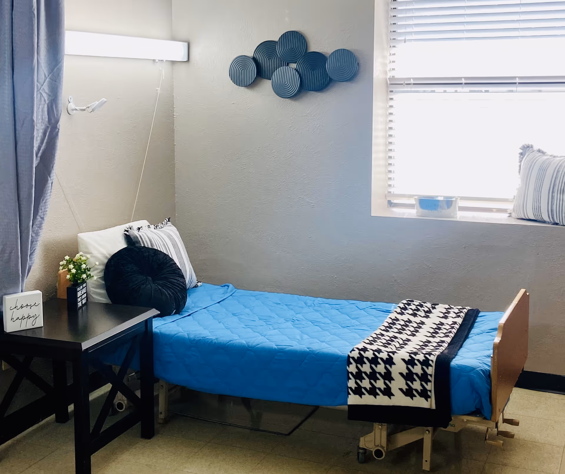 A single hospital-style bed with blue quilt, window, and bedside table in a bright room.