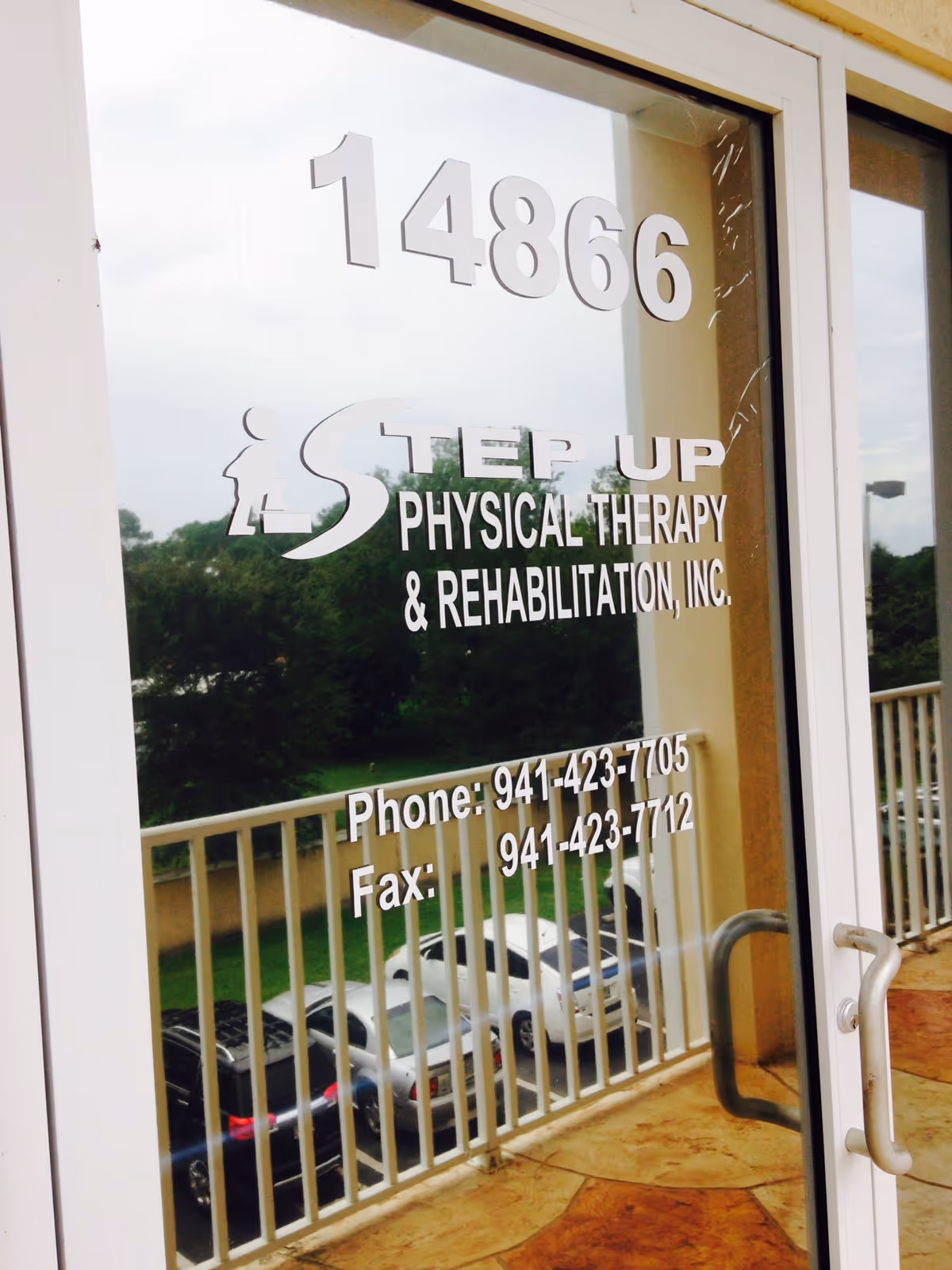 Glass door with Step Up Physical Therapy signage, address numbers, and parked cars outside.