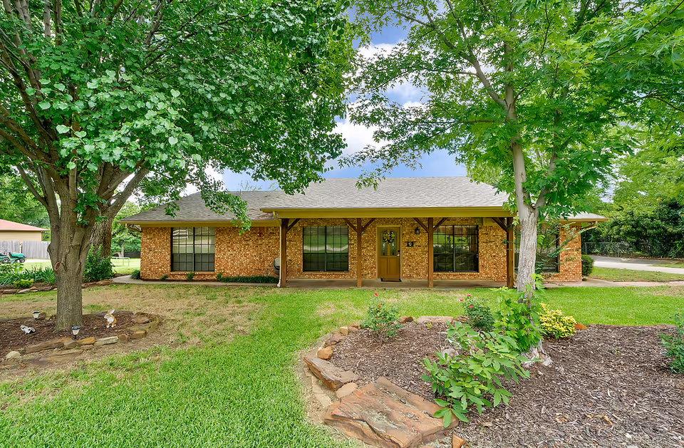 Front view of a brick senior living home with a porch, trees, and a green lawn.