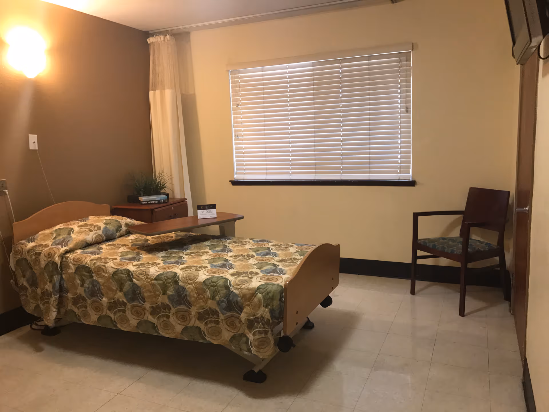 A simple bedroom in a care center featuring a single bed with a floral patterned bedspread, a small wooden bedside table with a plant and books, a wooden chair with a patterned cushion, a window with closed blinds, and a wall-mounted light fixture.