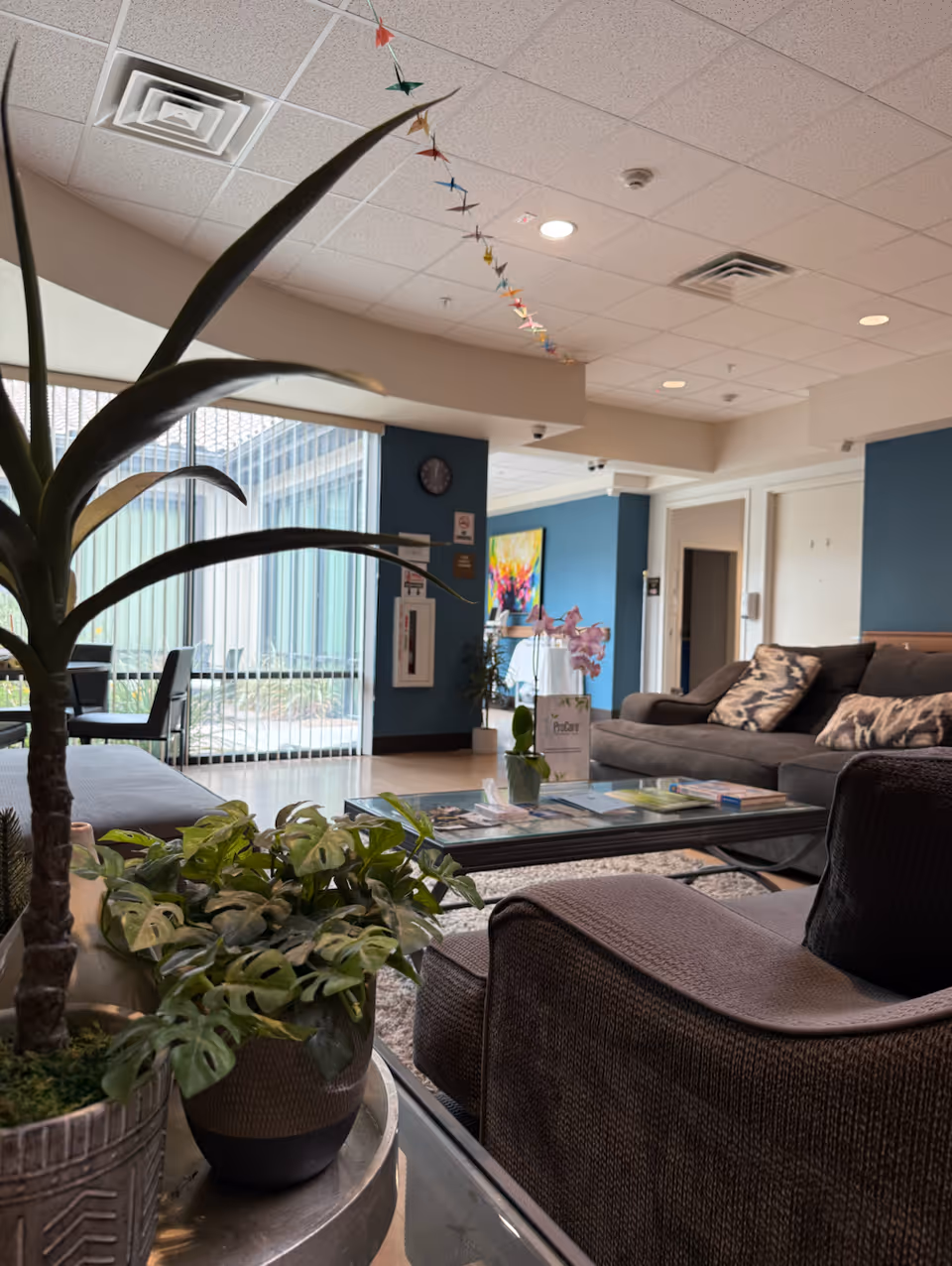 Interior senior living lounge with brown couches, glass coffee table, and potted plants.