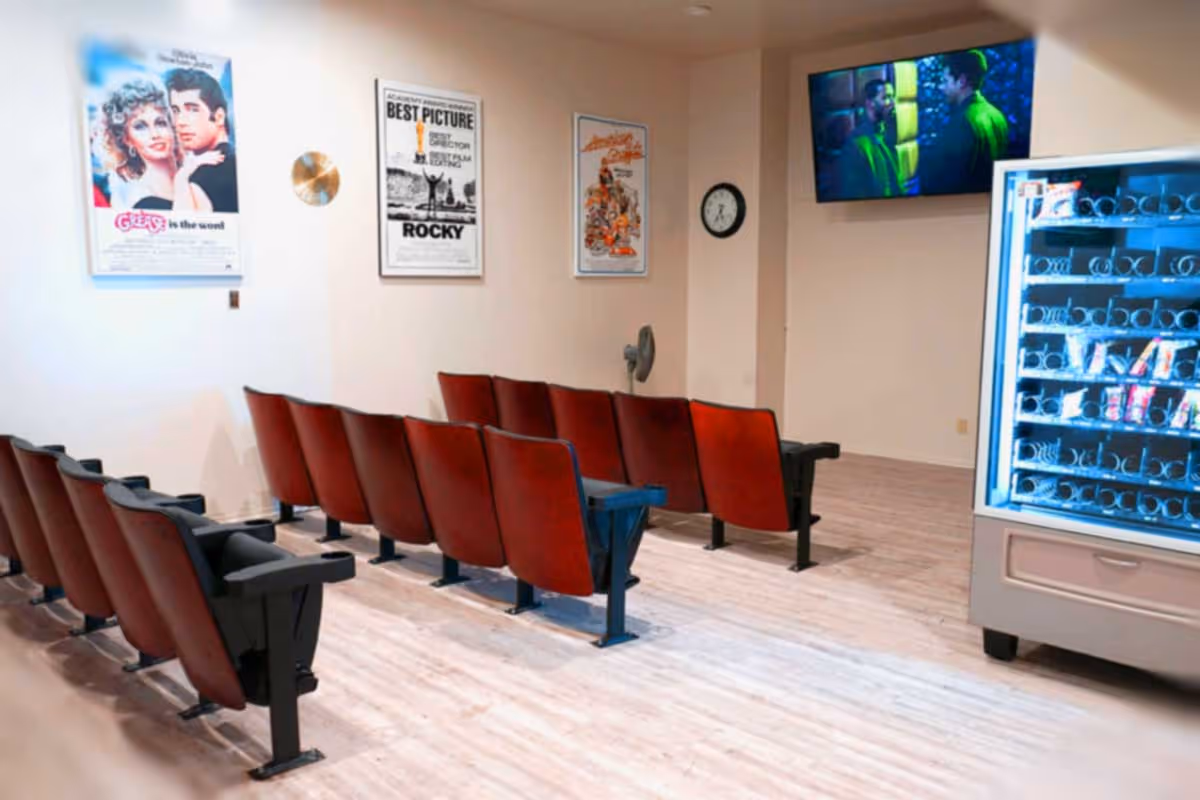 A small movie theater room with two rows of red theater seats facing a wall-mounted TV screen showing a movie scene. On the right side, there is a vending machine stocked with snacks. The walls have framed movie posters including Grease and Rocky, and a clock is mounted on the wall.