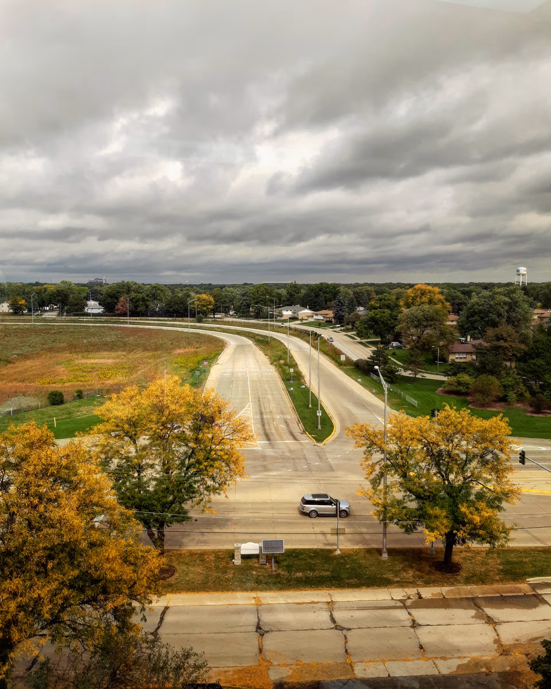 Aerial outdoor view of a curving road, autumn trees, and a cloudy sky near a facility.