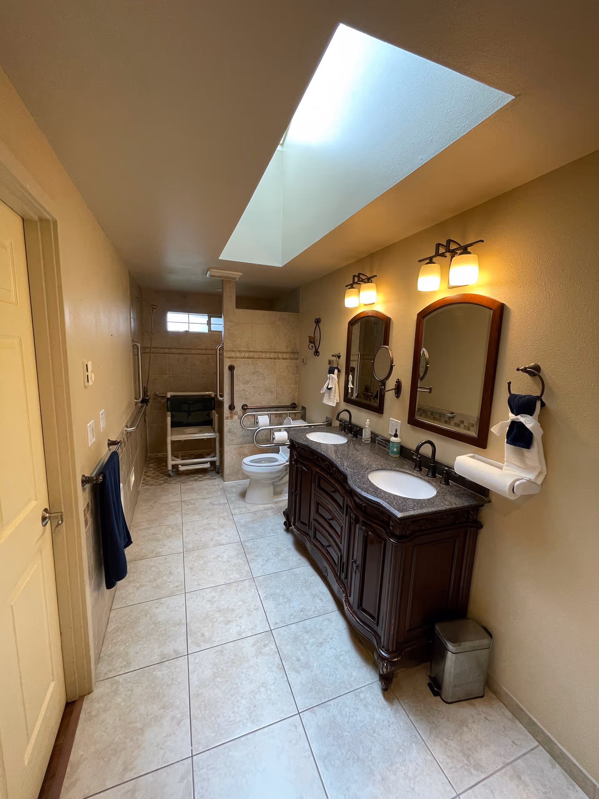 Accessible bathroom with double sink vanity, mirrors, wheelchair-accessible shower, toilet, skylight.