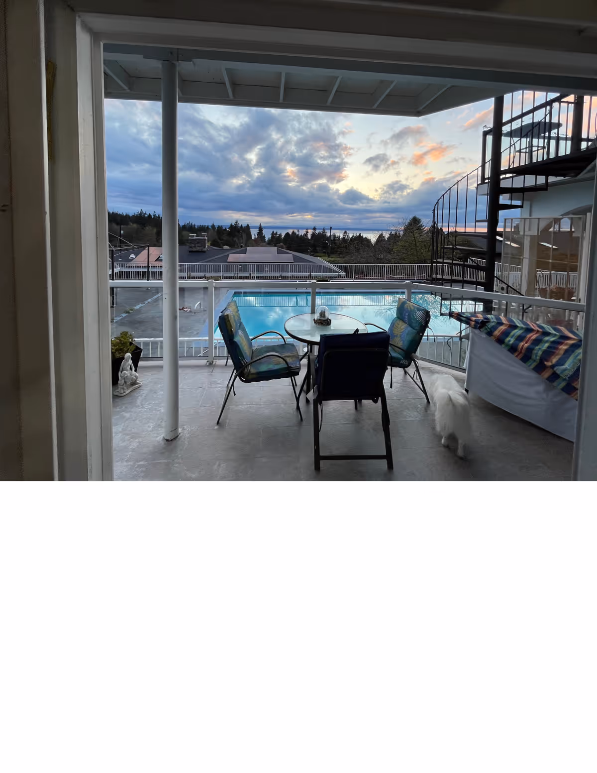Outdoor balcony with table and chairs overlooking a pool and sunset sky.