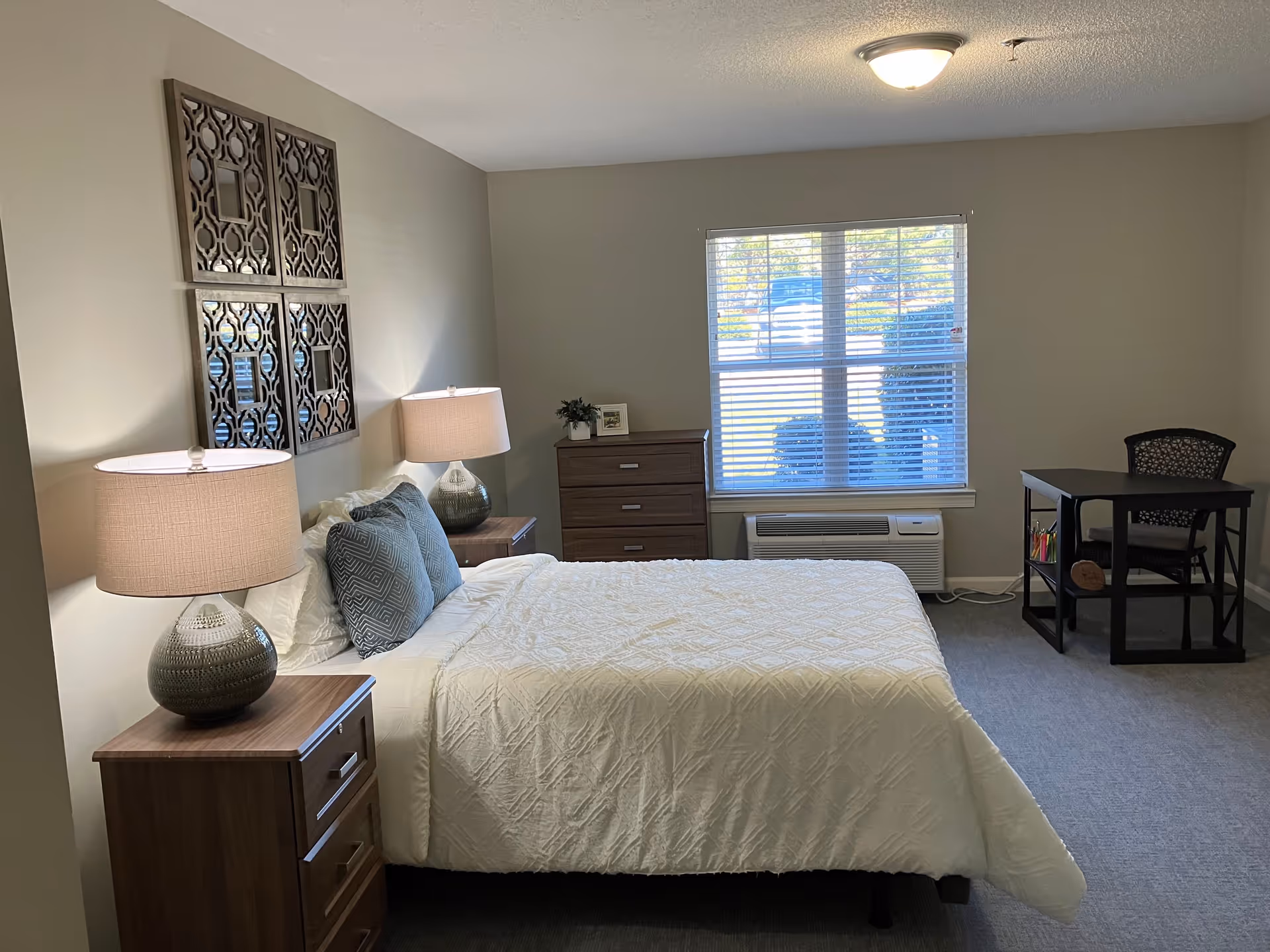 A furnished bedroom with a neatly made bed flanked by two lamps on nightstands, a dresser under a window, and a small desk in the corner.