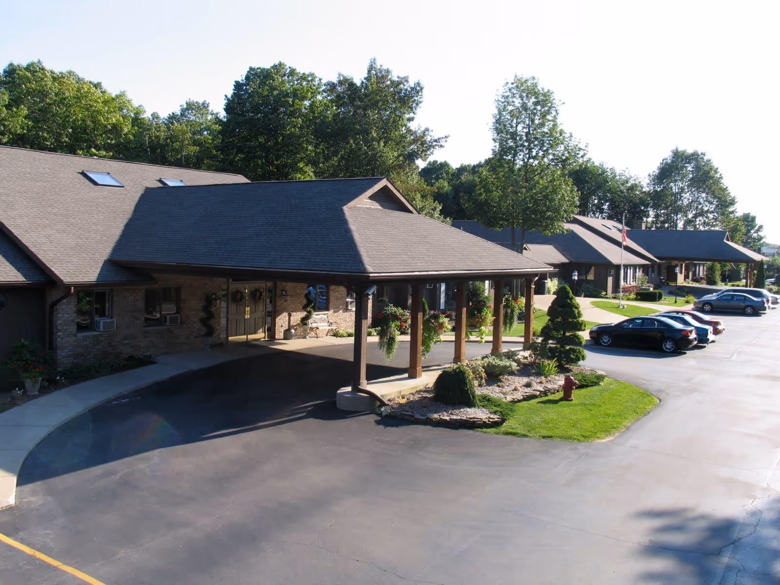 Front entrance of a single-story senior living facility with a covered porte-cochere, landscaped island, and parked cars.