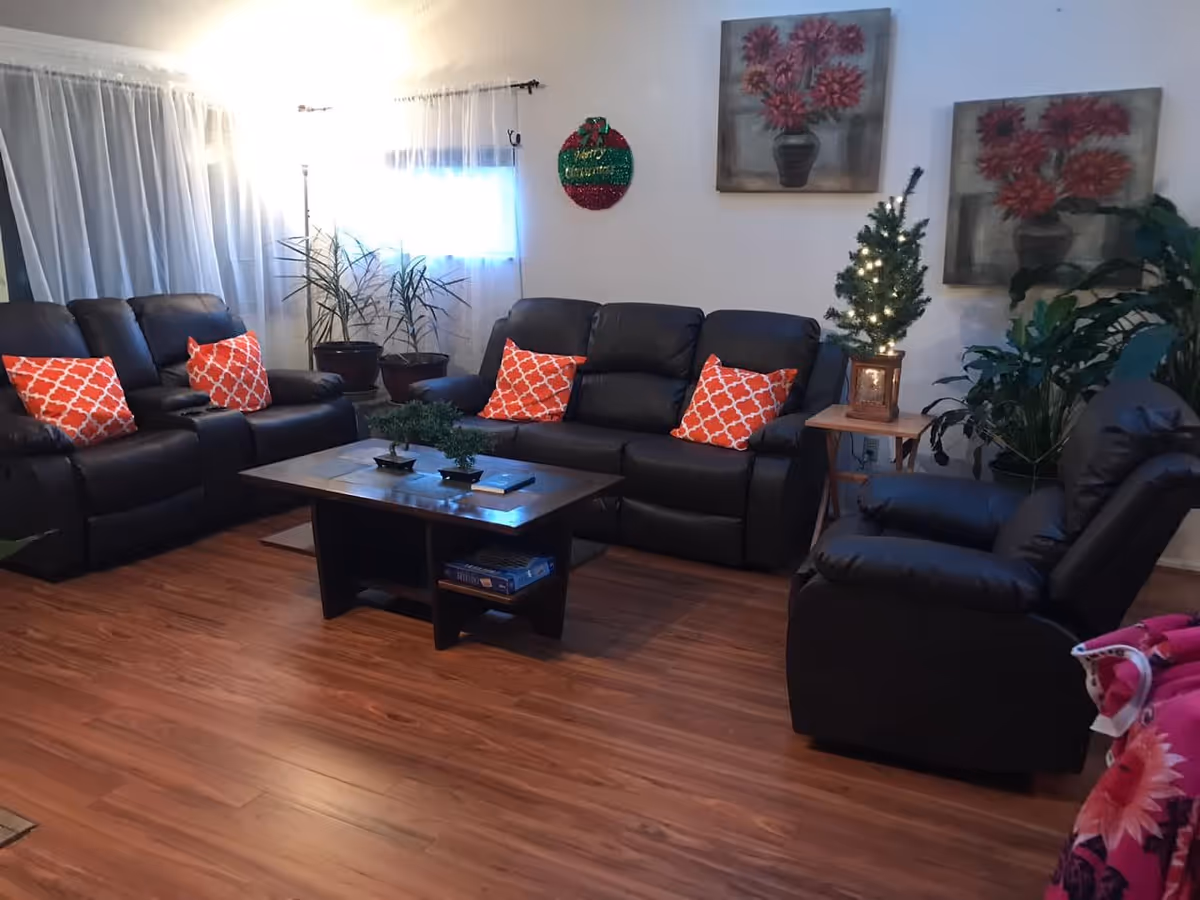 A cozy living room with dark leather sofas and armchairs, each adorned with orange patterned throw pillows. A wooden coffee table sits in the center with small plants on top. The room has wooden flooring, white walls with two paintings of red flowers, and a small decorated Christmas tree on a side table. There are also several potted plants near the window with sheer white curtains.
