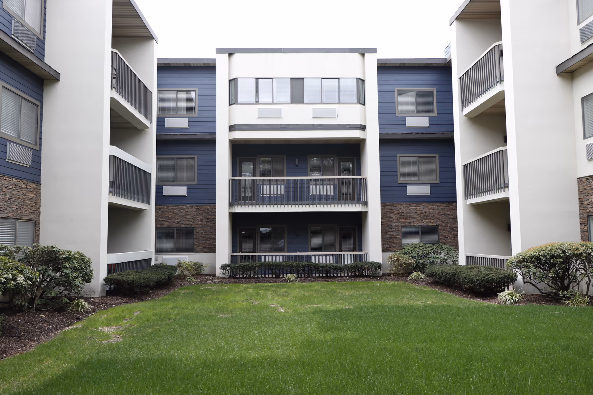 Green courtyard and lawn in front of a multi-story residential building with balconies and shrubs.