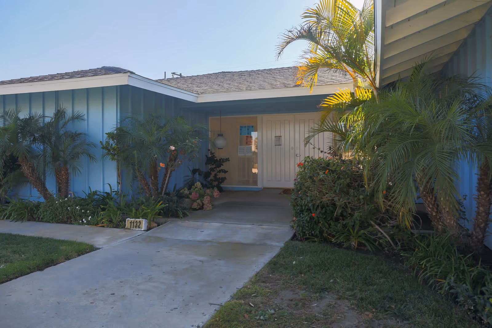 Entrance to a single-story building with blue exterior walls, a concrete pathway leading to double white doors, surrounded by green plants and palm trees under a clear blue sky.