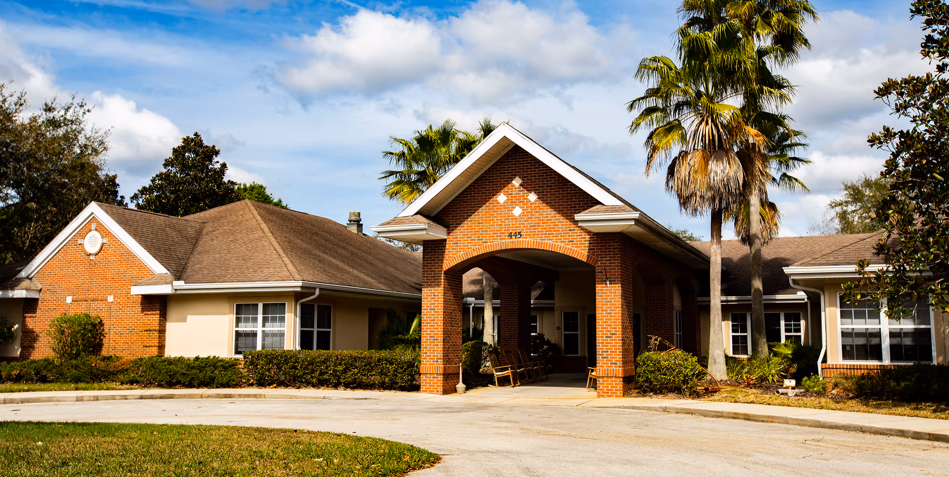 Brick senior living facility with a covered entrance, driveway, and palm trees against a blue sky.