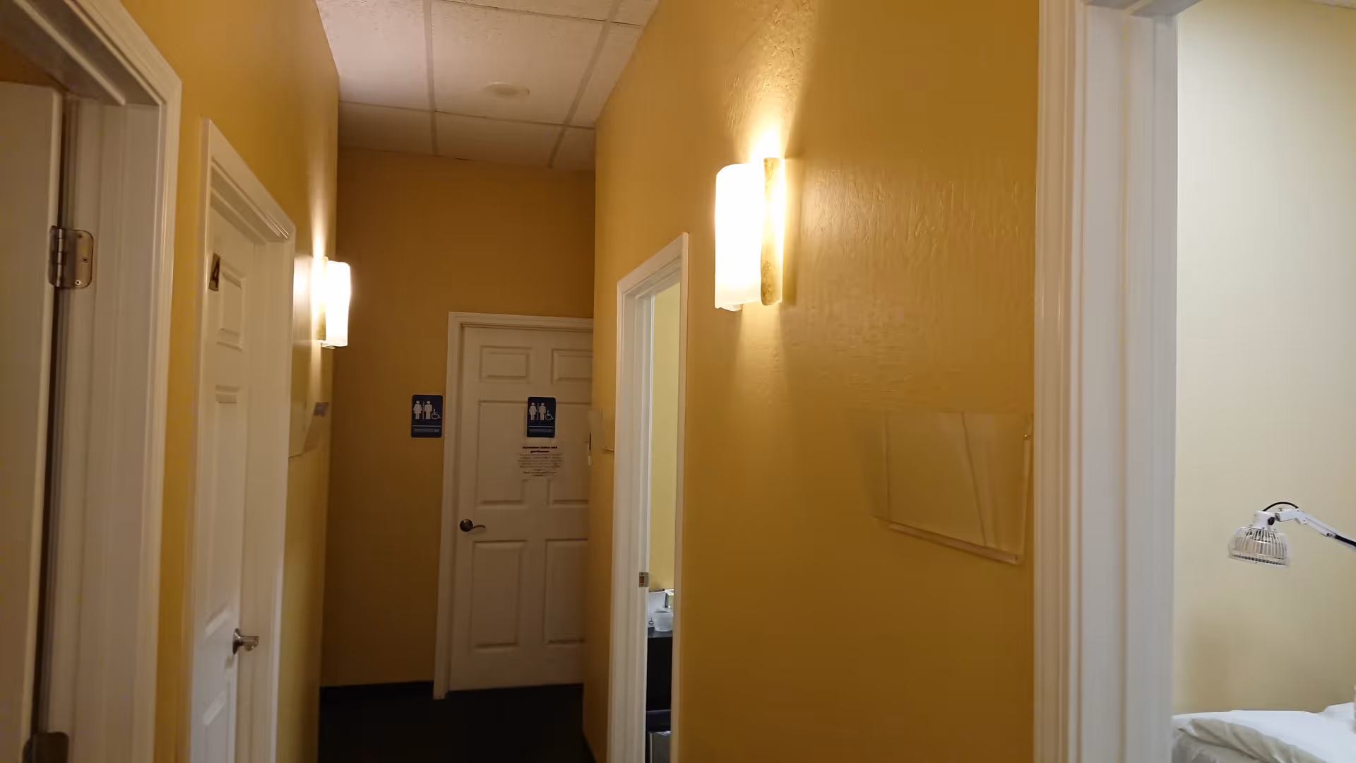 Yellow hallway inside a senior living facility with doors, wall sconces, and a visible patient room.