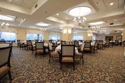 Spacious dining room with multiple tables covered in white tablecloths and surrounded by wooden chairs with cushioned seats. The room has patterned carpet flooring, large windows with floral valances, and several chandeliers hanging from the ceiling providing warm lighting.