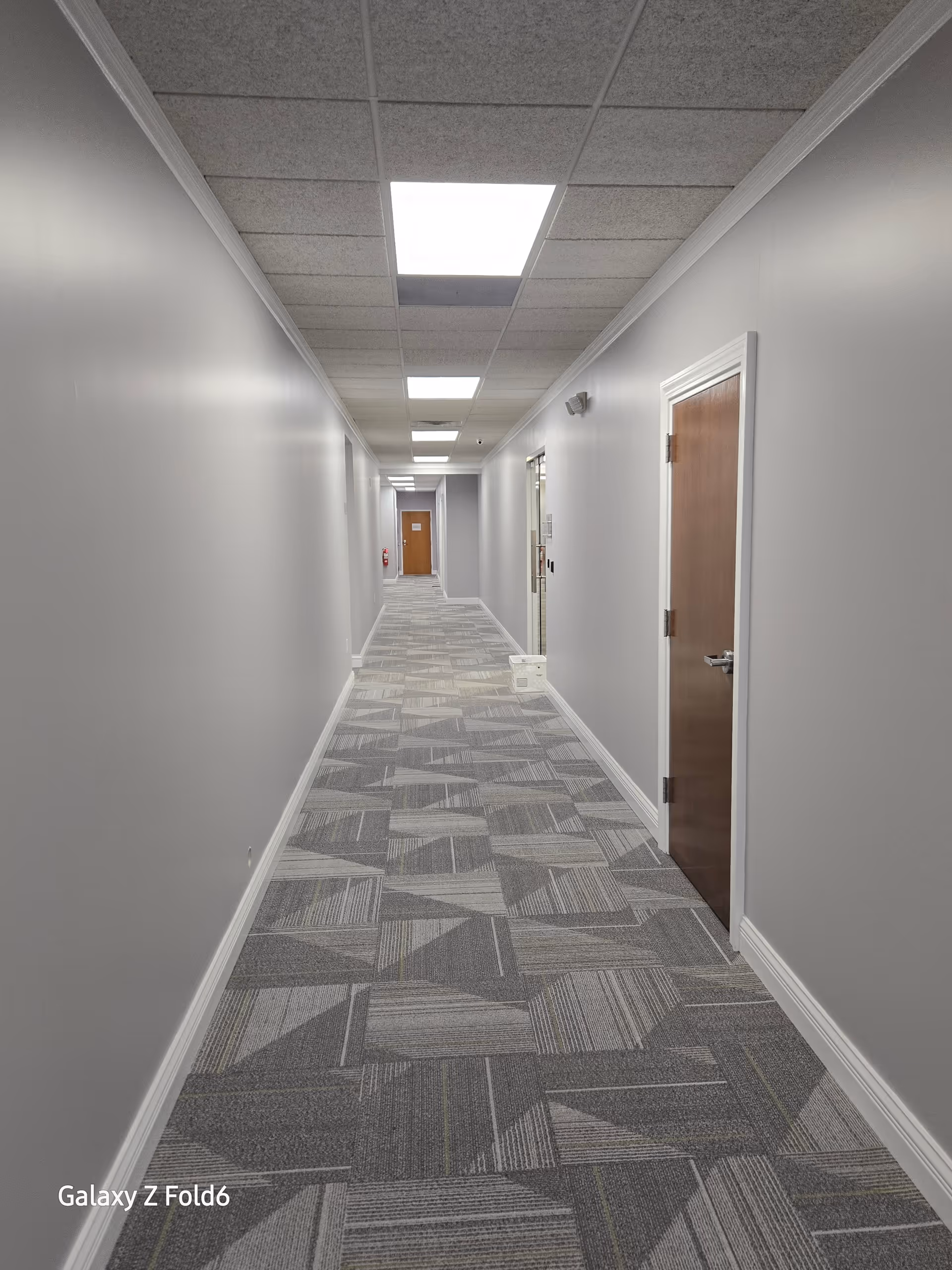 Long, well-lit interior hallway with doors along gray walls and patterned carpet.
