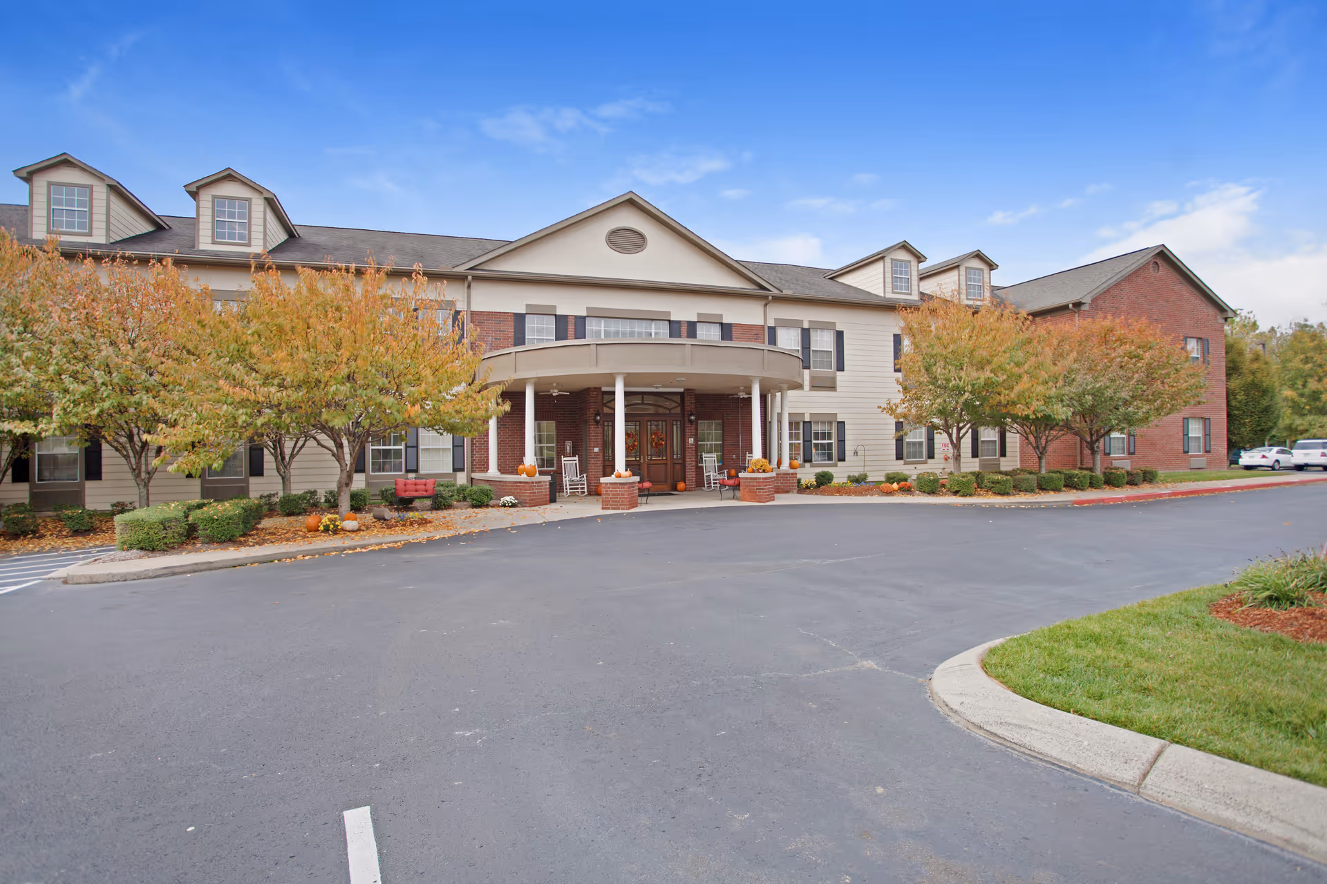 Exterior view of The Carriages of Rivergate senior living facility showing a two-story building with a covered entrance, surrounded by trees with autumn foliage and a paved driveway in front.