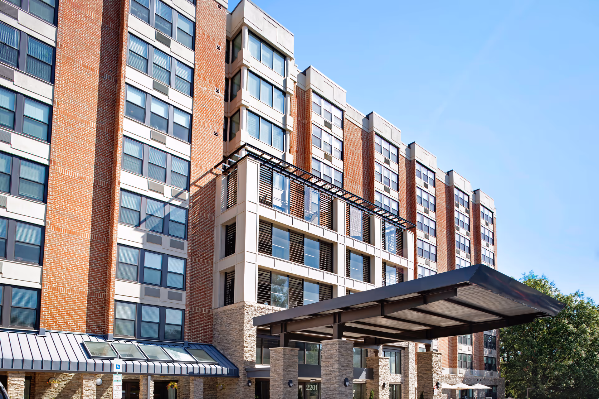 Multi-story brick senior living building with a covered entrance and glass windows under blue sky.