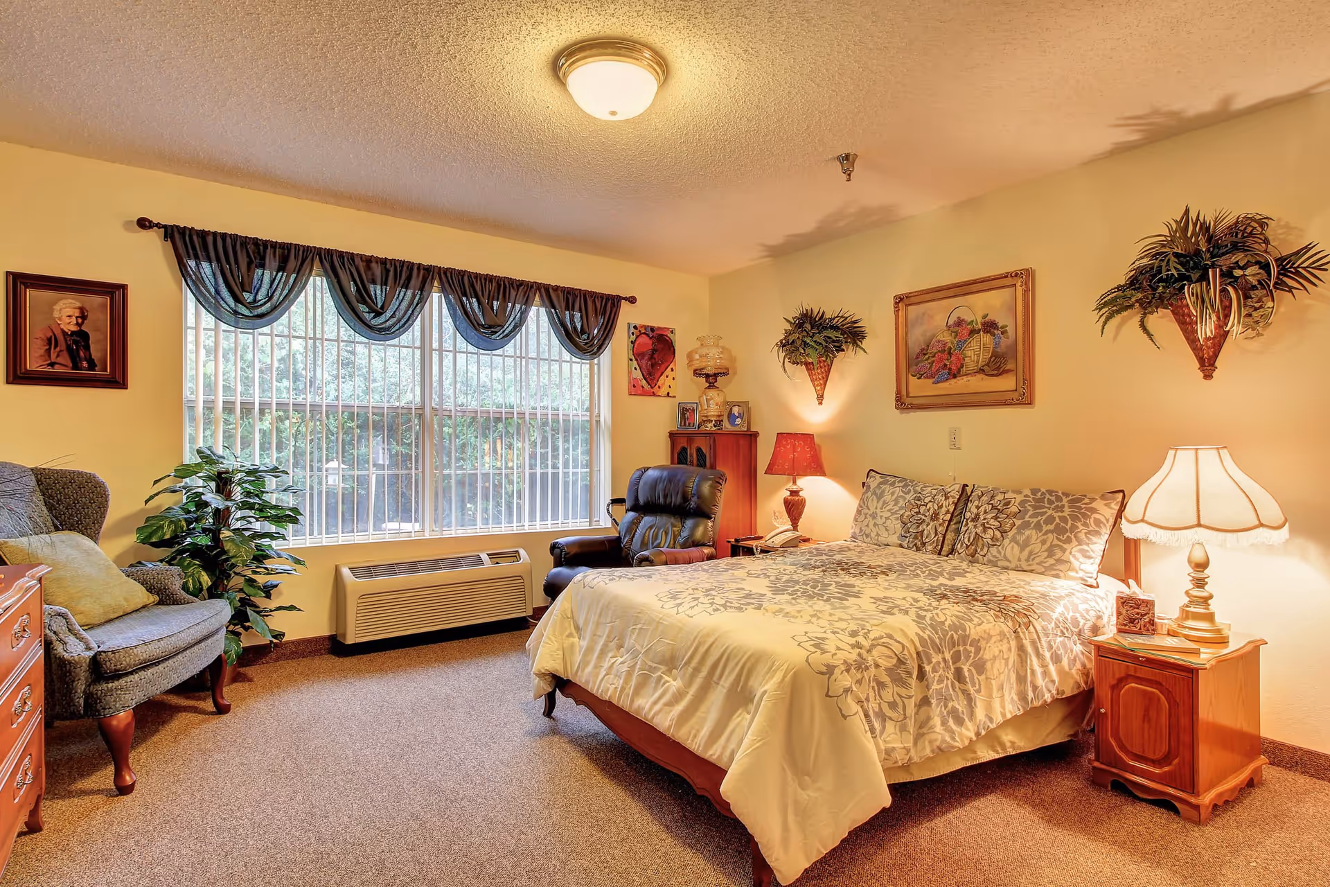 A warm, well-lit bedroom with a floral bed, bedside tables and lamps, an armchair and recliner, large window with drapes, and wall decorations.