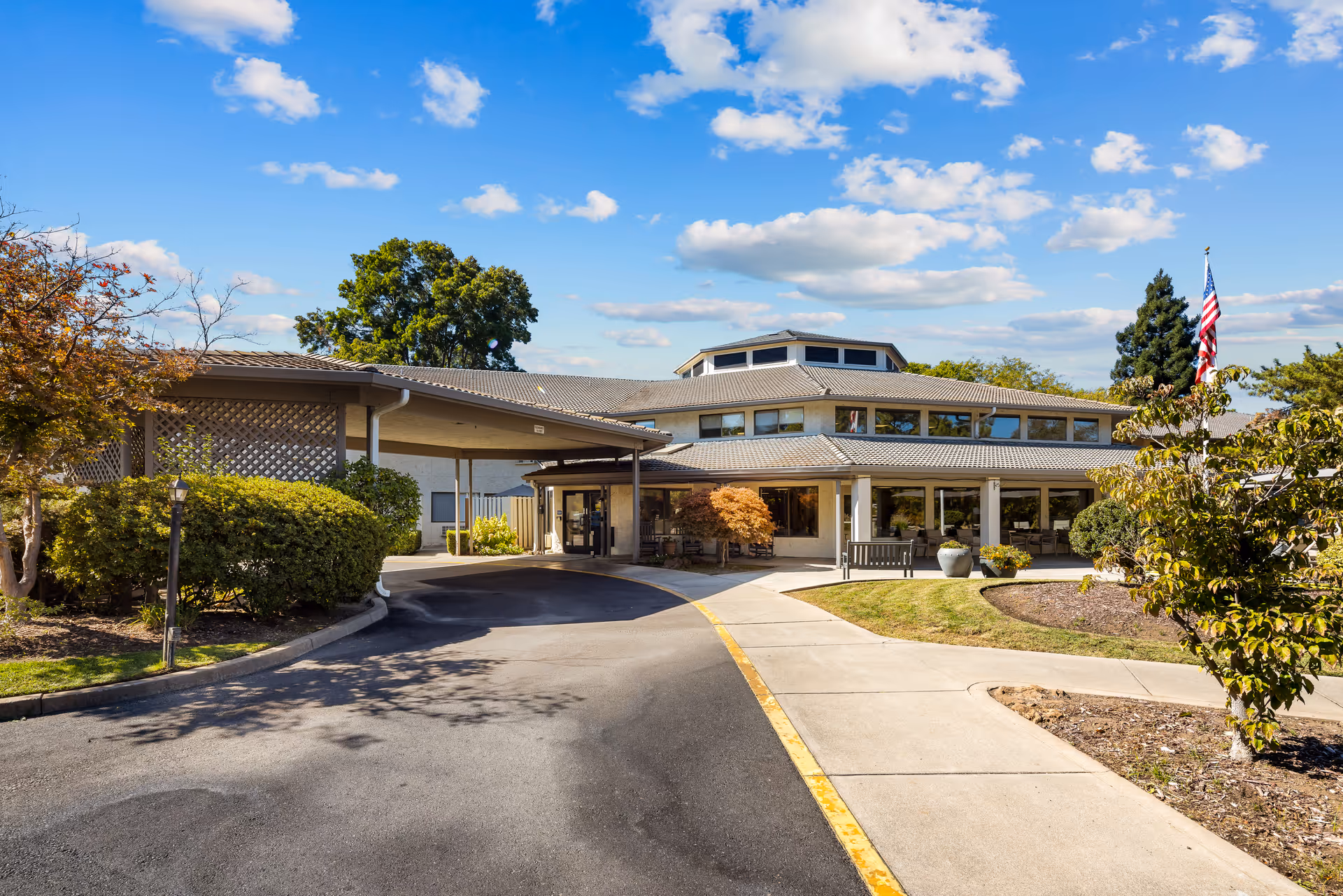 Senior living facility exterior with curved driveway, entrance, flag, and landscaped grounds.