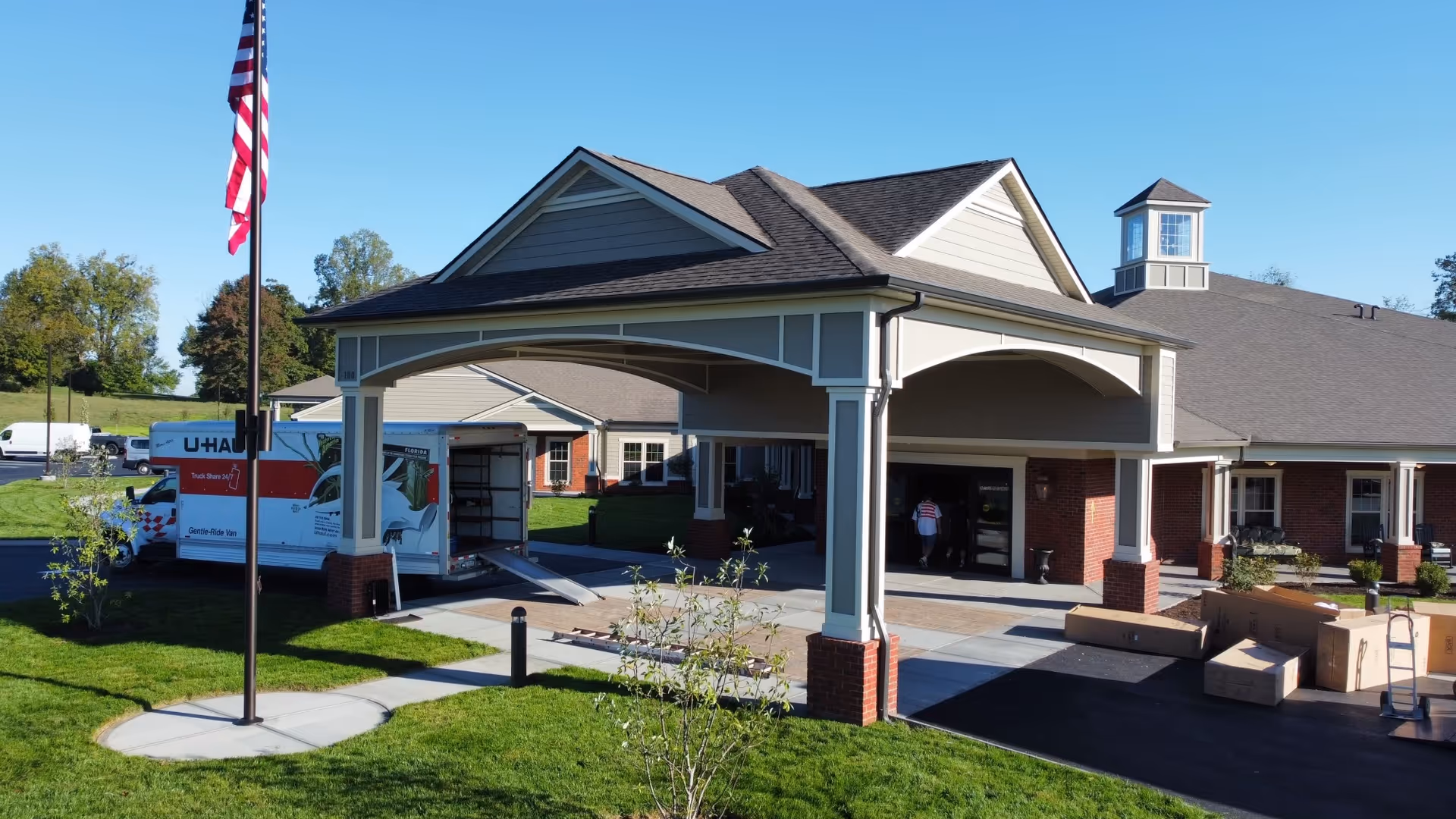 Exterior view of a senior living facility entrance with a covered drop-off area. A U-Haul truck is parked nearby with its back door open and a ramp extended. Several cardboard boxes and a hand truck are on the driveway. The building has a brick and siding facade with a cupola on the roof. An American flag is flying on a flagpole in the foreground.