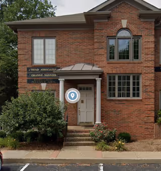 Brick building entrance with sign, white door, steps, and surrounding shrubs.